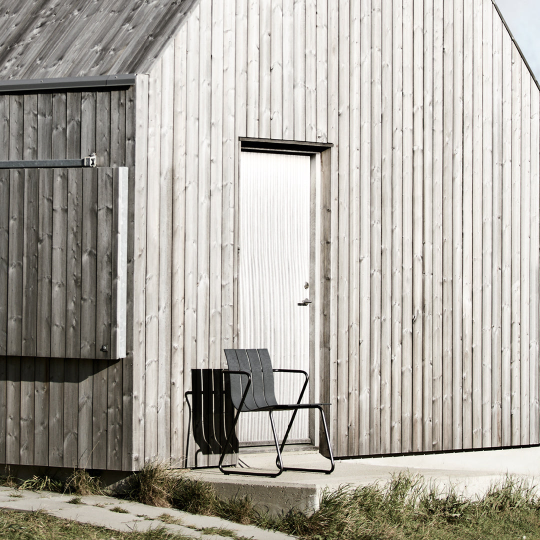 An Ocean Chair made of recycled material by Mater, in black, in front of a wood beach house.