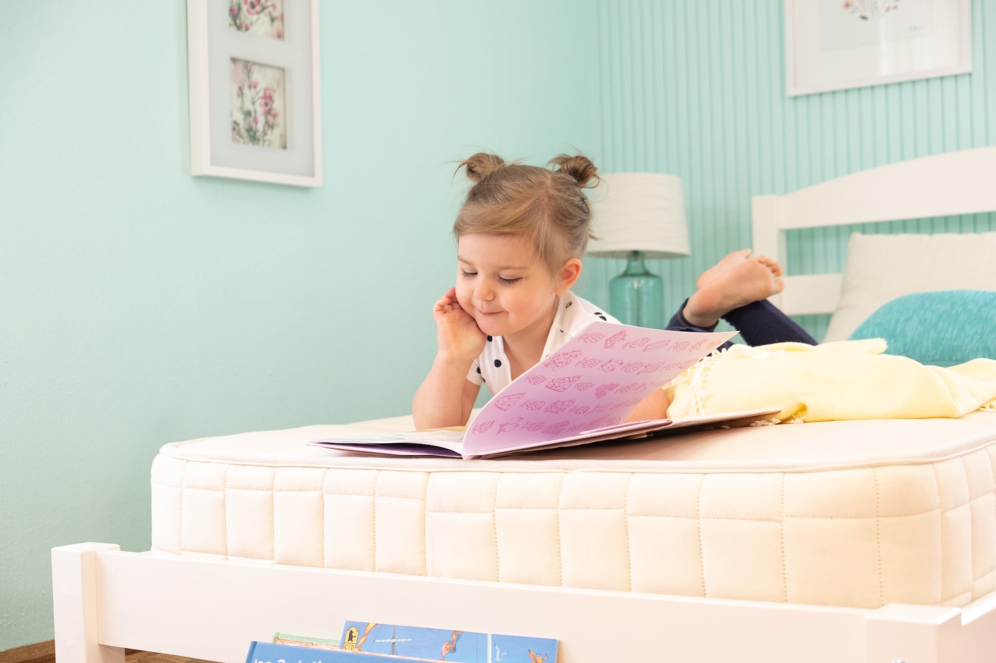 Young girl reading a book on a bed in a room with light blue walls and framed pictures.