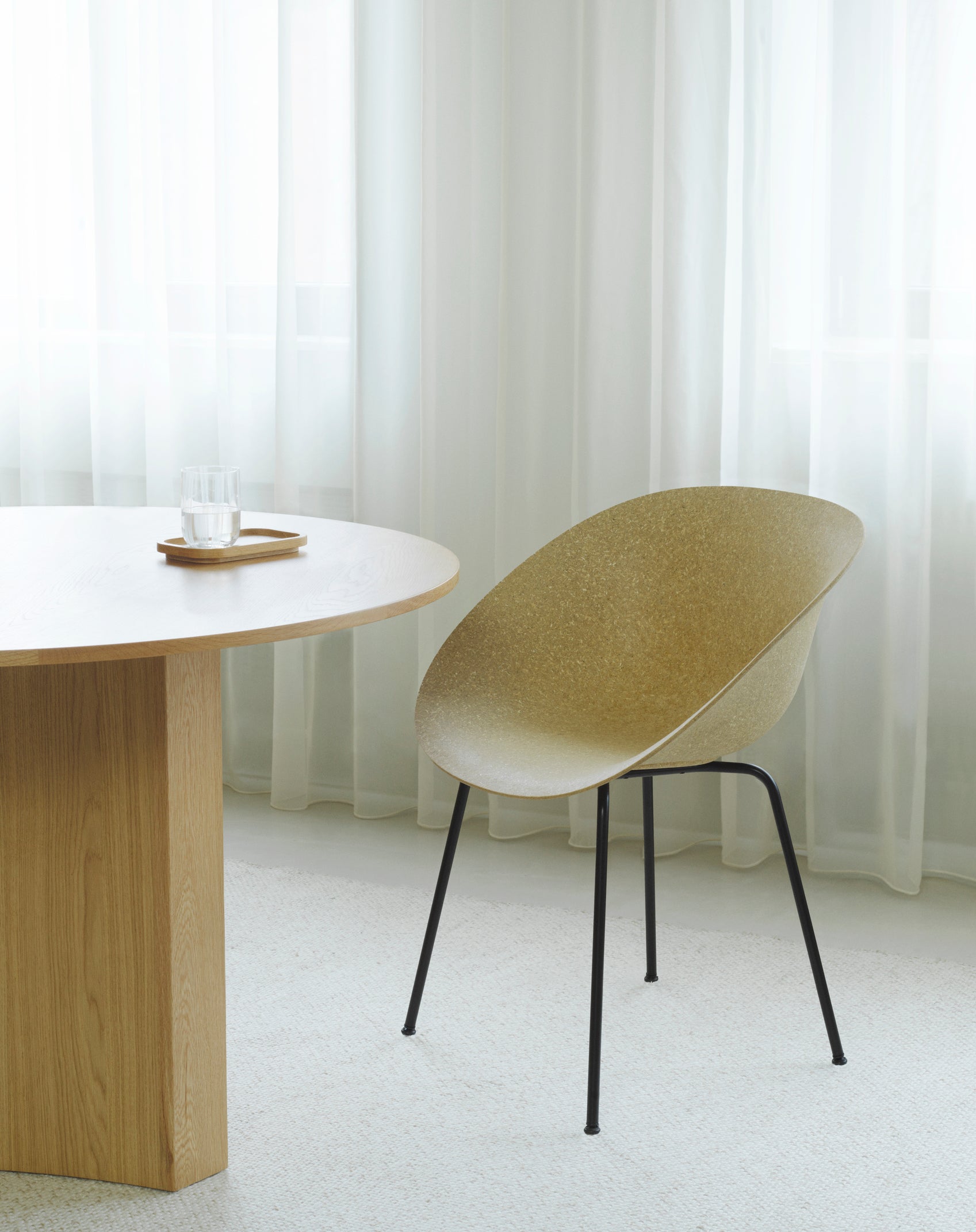 A sustainable armchair made with hemp plants by Normann Copenhagen, with black legs, at a light wood table with a glass of water on it.
