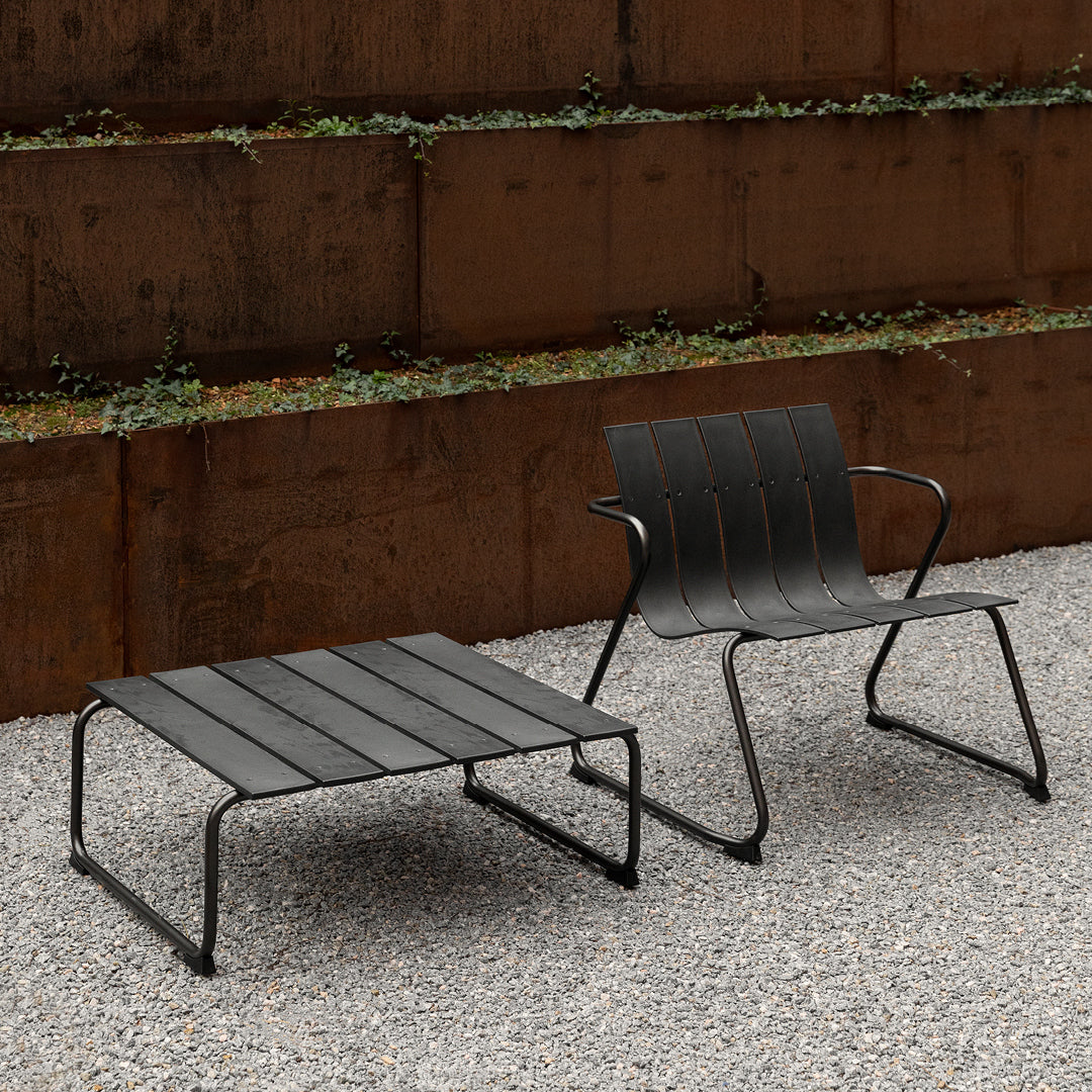 The Ocean Lounge Chair and Square Ocean Table in black, made of recycled fishing nets, next to a metal wall outdoors.