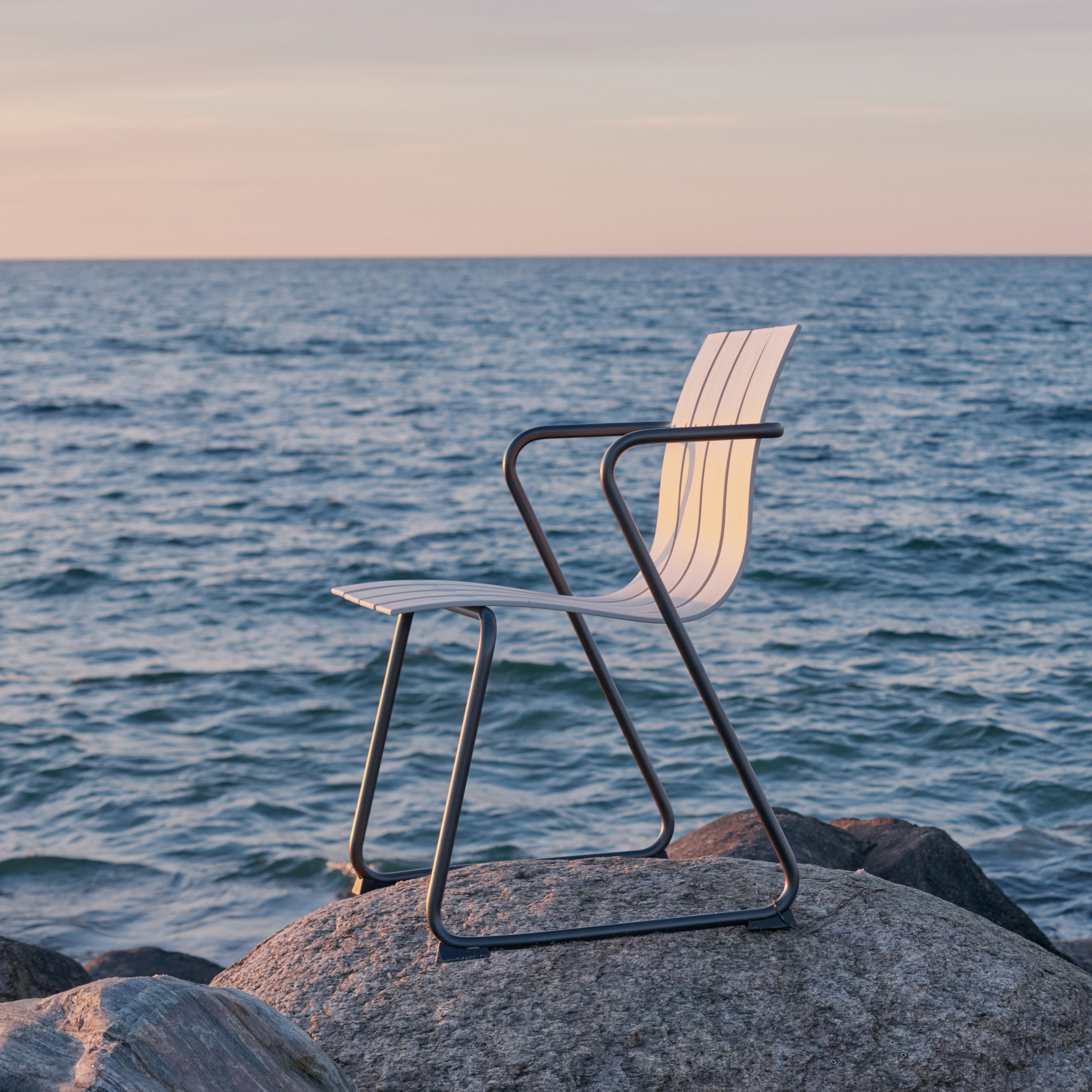 An Ocean Chair by Mater, made of recycled plastic fishing nets, in sand color, standing on a big rock, facing the ocean.